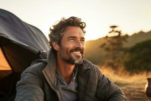 Portrait of man looking at camera while near camping tent at sunset AI generated photo