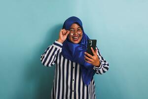 un emocionado de edad mediana asiático mujer en un azul hiyab, vistiendo los anteojos y un a rayas camisa, es riendo mientras mirando a el cámara y participación un móvil teléfono. ella es aislado en un azul antecedentes. foto