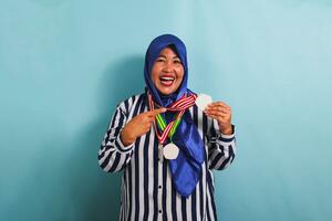 An excited middle-aged Asian woman, wearing a blue hijab and a striped shirt, is proudly pointing to the medal she is holding, isolated on blue background. photo