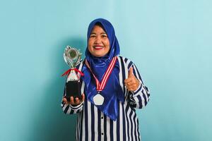 A happy middle-aged Asian businesswoman in a blue hijab and a striped shirt is showing an empty white medal while holding a silver trophy, celebrating her success, isolated on a blue background photo
