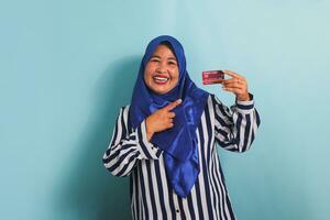 Excited Middle-aged Asian woman in blue hijab and striped shirt pointing finger at credit card, recommending payment method, going on shopping, isolated over blue background. photo