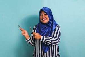 An excited middle-aged Asian woman in a blue hijab and a striped shirt is pointing to the left copyspace with a spoon and fork. She is isolated on a blue background photo