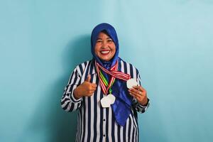 An excited middle-aged Asian woman in a blue hijab and a striped shirt is giving a thumbs-up while holding a medal, isolated on a blue background photo