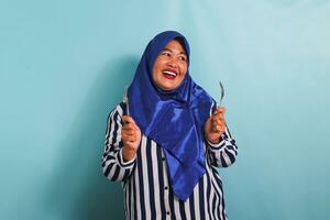 An excited middle-aged Asian woman in a blue hijab and a striped shirt is holding a spoon and fork, feeling hungry while waiting for food to be served. She is isolated on a blue background photo