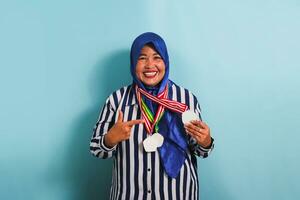 un emocionado de edad mediana asiático mujer, vistiendo un azul hijab y un a rayas camisa, es con orgullo señalando a el medalla ella es tenencia, aislado en azul antecedentes. foto