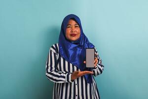 An Excited middle-aged Asian woman in a blue hijab and a striped shirt is showing a phone with a blank white screen. She is isolated on a blue background photo