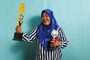 An excited middle-aged Asian businesswoman in a blue hijab and a striped shirt is holding a gold and silver trophy, celebrating her success and achievement. She is isolated on a blue background photo