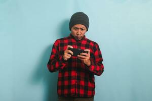 Confused young Asian man with a beanie hat and a red plaid flannel shirt feels disappointed and annoyed after losing while playing an online game on his smartphone, isolated on a blue background photo