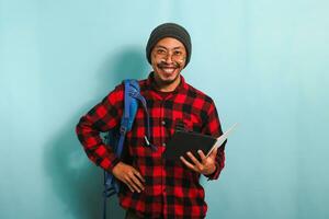 joven asiático estudiante hombre vistiendo un mochila es participación libros mientras en pie en contra un azul antecedentes foto