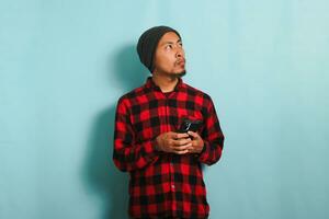 Pensive young Asian man with a beanie hat and a red plaid flannel shirt is deep in thought, looking at copy space, holding his phone as he considers and makes a decision, isolated on a blue background photo