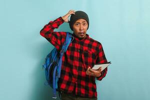 A surprised young Asian student with a beanie hat and a red plaid flannel shirt, wearing a backpack, scratching his head appears unhappy and confused, isolated on a blue background photo