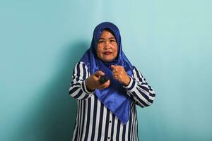 An annoyed middle-aged Asian woman in a blue hijab and a striped shirt is trying to switch channels while watching a movie on TV. She is isolated on a blue background photo