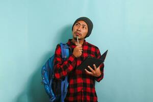 Pensive young Asian student is deep in thought, looking ideas while standing against blue background photo