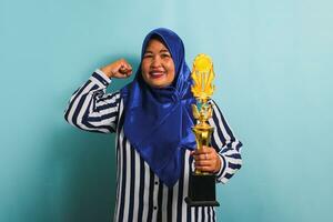 un orgulloso de edad mediana asiático mujer de negocios en un azul hijab y un a rayas camisa es haciendo un fuerte gesto mientras participación un oro trofeo, celebrando su éxito. ella es aislado en un azul antecedentes. foto