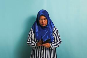 Speechless middle-aged Asian woman in blue hijab and striped shirt is looking into her empty wallet with a frustrated expression, isolated on a blue background. Bankruptcy, financial crisis concept. photo