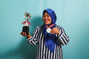 un contento de edad mediana asiático mujer de negocios en un azul hijab y un a rayas camisa es demostración un vacío blanco medalla mientras participación un plata trofeo, celebrando su éxito, aislado en un azul antecedentes foto