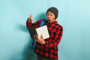 Excited young Asian student is pleasantly surprised by a book he read, isolated on blue background photo