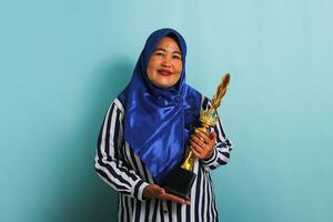 un emocionado de edad mediana asiático mujer de negocios en un azul hijab y un a rayas camisa es participación un oro trofeo, celebrando su éxito y logro. ella es aislado en un azul antecedentes foto