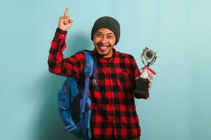 Excited Young Asian man student proudly pointing a trophy in his hand, isolated on blue background photo