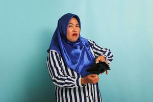 Unhappy middle-aged Asian woman in hijab and striped shirt is showing her empty wallet to the camera with a frustrated expression, isolated on a blue background. Bankruptcy, financial crisis concept. photo
