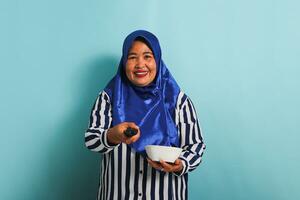 An excited middle-aged Asian woman in a blue hijab and a striped shirt is holding a white bowl while turning up the TV volume and watching an interesting TV show. She is isolated on a blue background photo