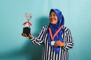 A happy middle-aged Asian businesswoman in a blue hijab and a striped shirt is showing an empty white medal while holding a silver trophy, celebrating her success, isolated on a blue background photo