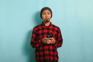 An upset Young Asian man with a beanie hat and a red plaid flannel shirt is looking at camera holding a mobile phone, experiencing problems and feeling angry and annoyed, isolated on a blue background photo