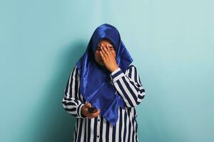 An anxious middle-aged Asian woman in a blue hijab and a striped shirt is touching her head, feeling stressed, while holding a TV remote. She is isolated on a blue background photo