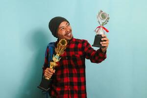 Happy young Asian man student lifting up his trophies, isolated on blue background. photo