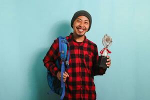 Excited Young Asian man student holding a trophy, smiling at the camera, isolated on blue background photo