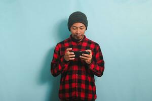 Excited young Asian man with a beanie hat and a red plaid flannel shirt is playing an online game on his smartphone, isolated on a blue background photo