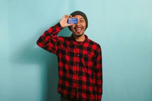 Joyful young Asian man is playfully covering one eye with a credit card , isolated on blue backdrop photo