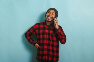 Happy Young Asian man with beanie hat and red plaid flannel shirt is laughing while talking on his smartphone, having a good conversation, hearing funny jokes, isolated on a blue background photo