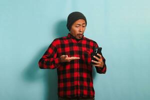 Funny Young Asian man with a beanie hat and a red plaid flannel shirt is in shock and surprised while reading bad news on his smartphone, isolated on a blue background. photo