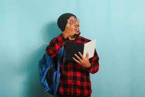 Excited young Asian student with a beanie hat and a red plaid flannel shirt, wearing a backpack and glasses, is holding books and pointing aside the copy space while standing against a blue background photo