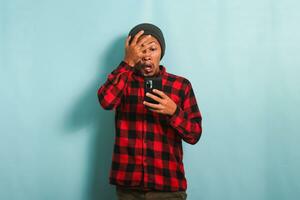 Funny Young Asian man with beanie hat and red plaid shirt is peeking at inappropriate content on his phone, shocked and covering his face, looking through his fingers, isolated on blue backdrop photo