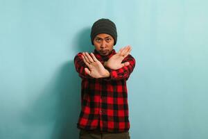 Angry young Asian man stands with his arms crossed, stop gesture, isolated on blue background. photo