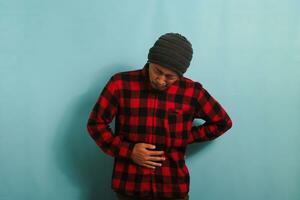 Unhappy young Asian man is suffering from a stomach ache, bending over, isolated on blue background photo