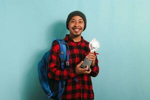 Excited young Asian man student makes YES gesture while holding trophy, isolated on blue background photo