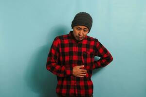 Unhappy young Asian man is suffering from a stomach ache, bending over, isolated on blue background photo