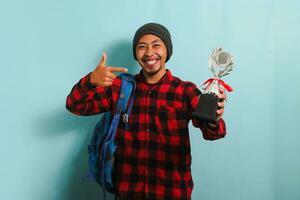 Excited Young Asian man student proudly pointing a trophy in his hand, isolated on blue background photo