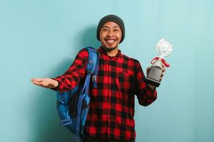 emocionado joven asiático hombre estudiante participación un trofeo, sonriente a el cámara, aislado en azul antecedentes foto