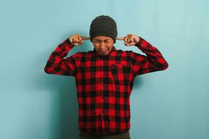 Annoyed young Asian man with a beanie hat and a red plaid flannel shirt covers his ears, feeling the painful ache of an earache and suffering from otitis due to loud noise, isolated on blue background photo