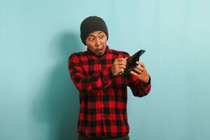 Upset young Asian man with beanie hat and red plaid flannel shirt showing an empty wallet to the camera, isolated on a blue background. Depicting bankruptcy, inflation, and financial crisis concept photo
