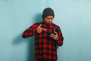 Angry young Asian man with a beanie hat and a red plaid flannel shirt is yelling and pointing at his phone, isolated on a blue background photo