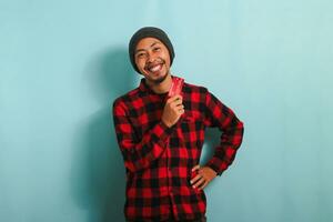 Excited young Asian man with a beanie hat and red plaid flannel shirt is holding a bank credit card in his hands, ready to make a purchase in an online store while standing against a blue background photo