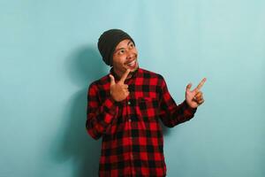 Excited Young Asian man with a beanie hat and a red plaid flannel shirt is pointing his finger to the right at the copy space, isolated on a blue background photo