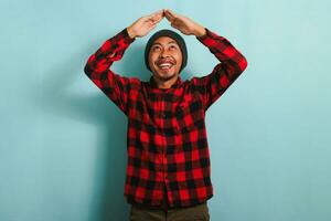 Happy young Asian man with a beanie hat and red plaid flannel shirt making a roof gesture above his head with a beaming smile, isolated on a blue background photo
