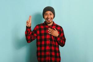 A young Asian man with a beanie hat and red plaid flannel shirt is swearing with his hand on his chest and an open palm, making a loyalty promise oath, isolated on a blue background photo