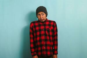 Displeased young Asian man with a beanie hat and a red plaid flannel shirt looks aside at an empty copyspace with a disgusted expression while standing against a blue background photo